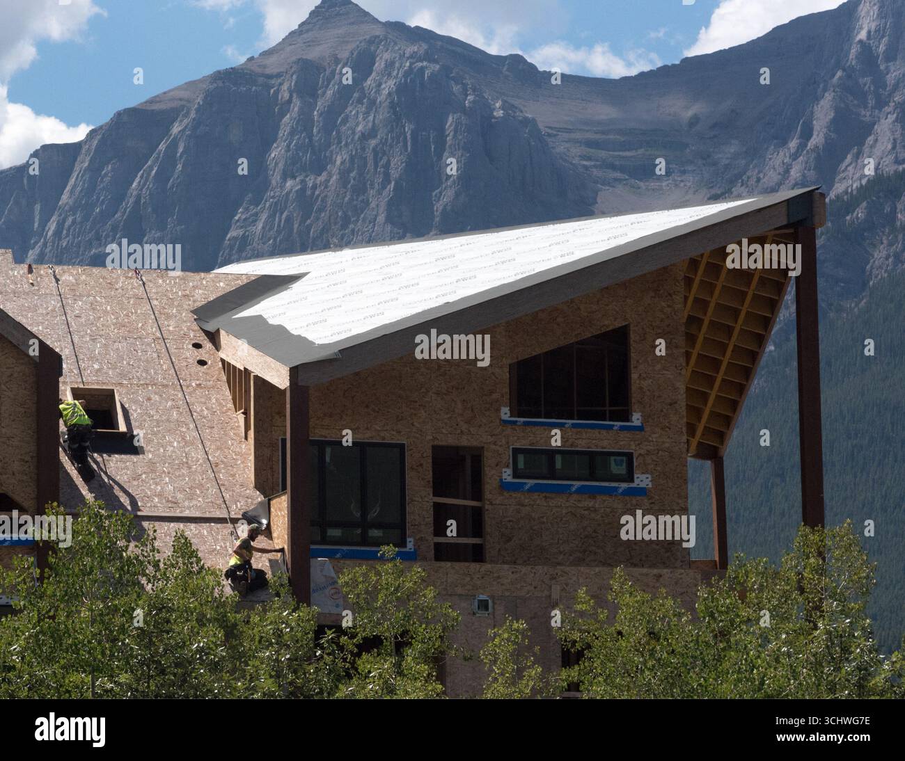Zwei Bauarbeiter, die Kabelbäume verwenden, installieren Dachmaterialien in einem neuen Wohngebäude in Canmore, Alberta. Rundle Mountain bietet eine Kulisse. Stockfoto