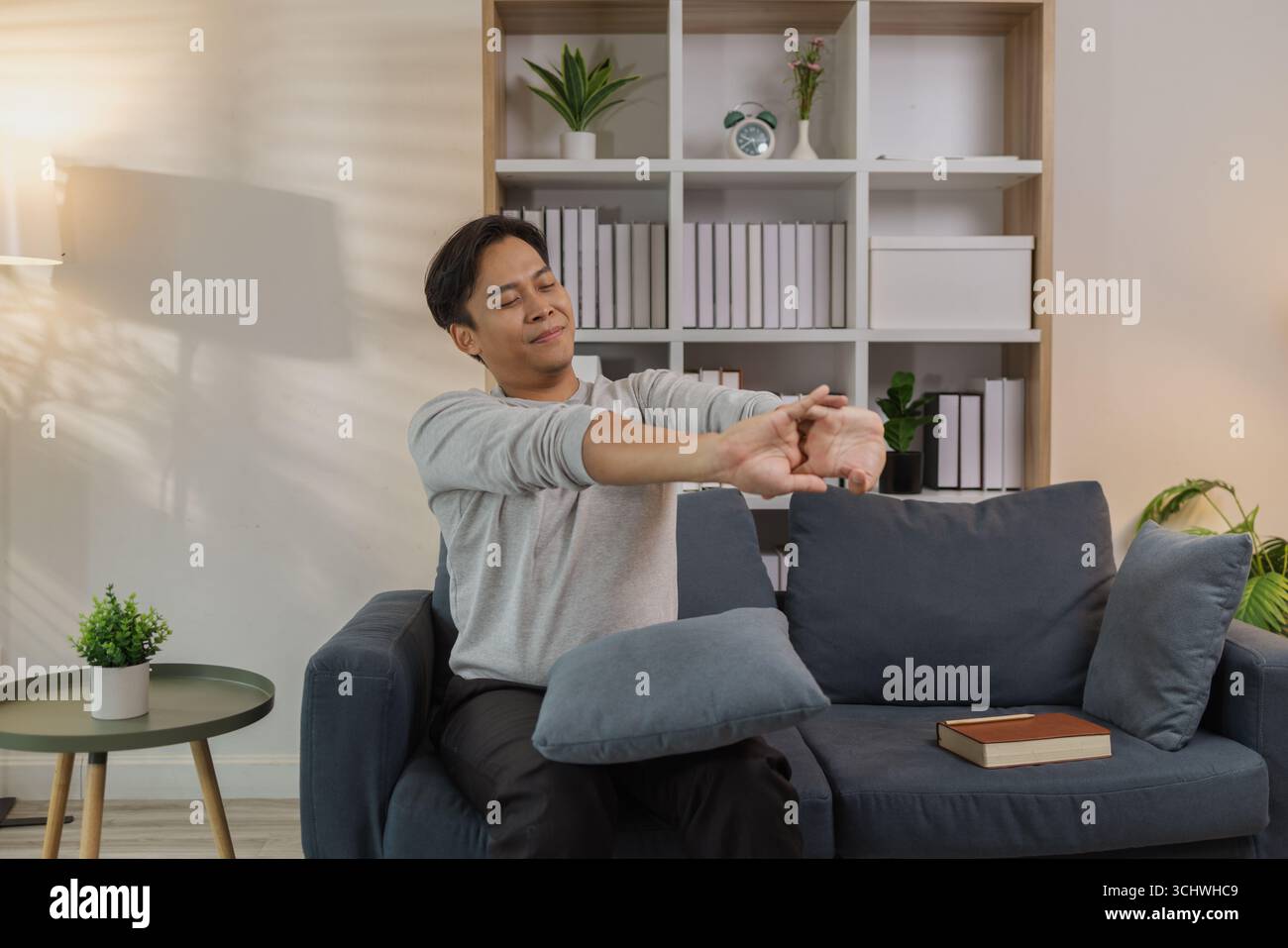 Ein junger Mann in einem grauen Hemd entspannt sich und dehnt seine Arme bequem, während er zu Hause auf einem gemütlichen blauen Sofa sitzt. Er genießt einen friedlichen Moment, lächelt Stockfoto