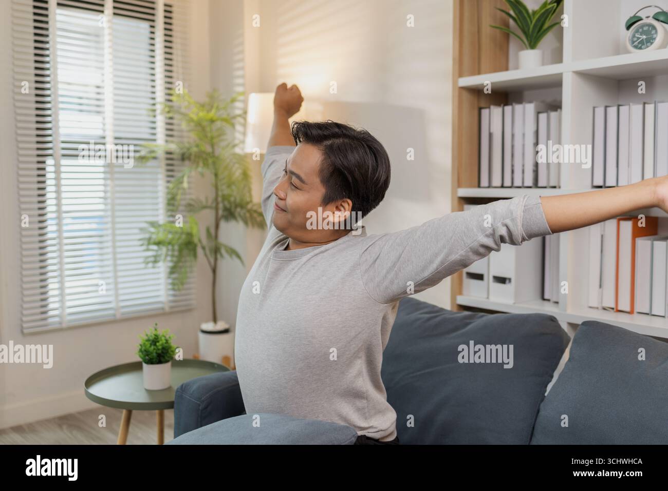Ein junger Mann in einem grauen Hemd entspannt sich und dehnt seine Arme bequem, während er zu Hause auf einem gemütlichen blauen Sofa sitzt. Er genießt einen friedlichen Moment, lächelt Stockfoto