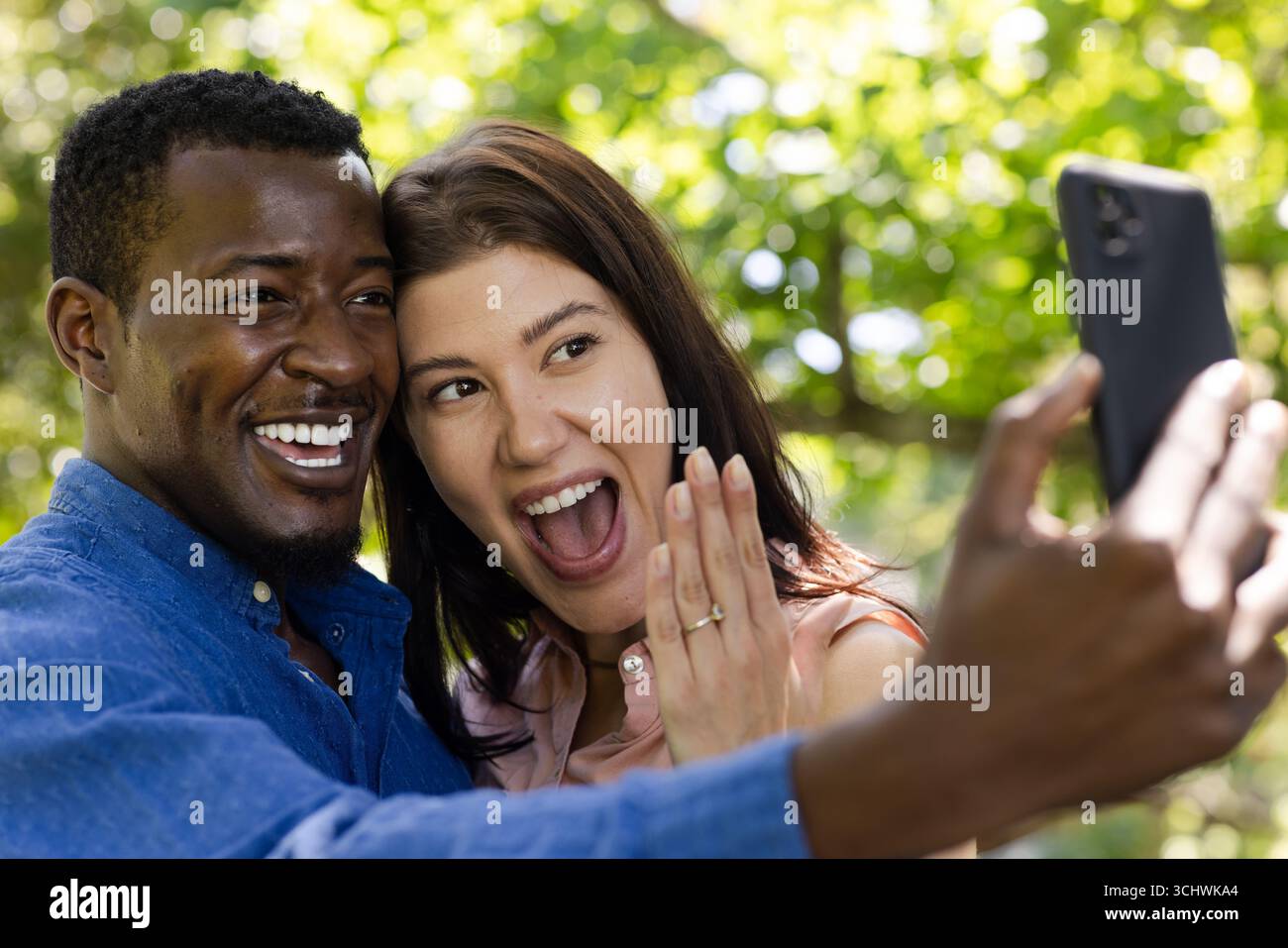 Fröhliches, vielseitiges Paar, das im Freien Selfie macht und mit großem Lächeln die Verlobung feiert Stockfoto