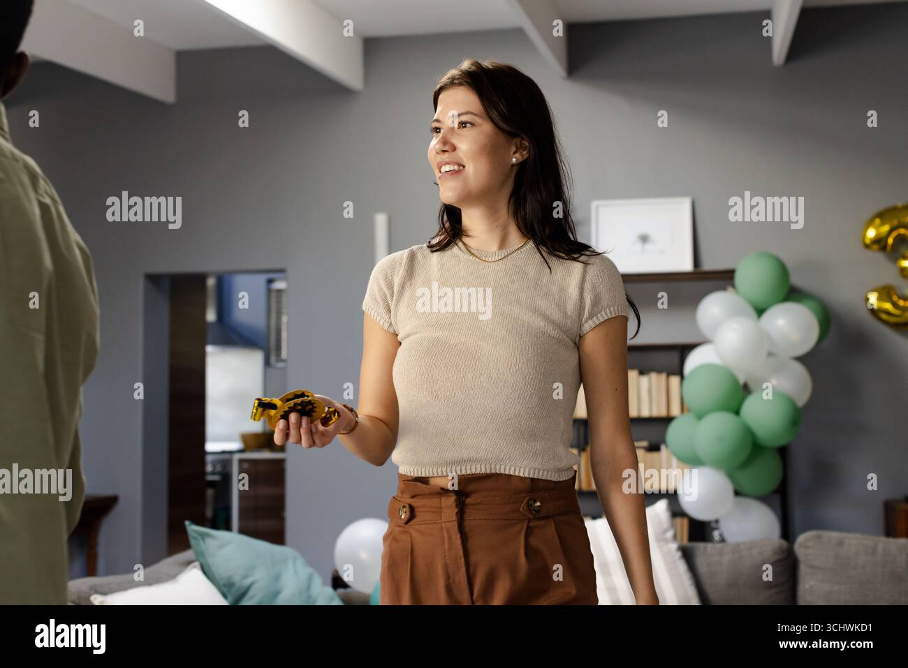 Frau, die Partyfavorit hält, lächelt im Wohnzimmer mit festlichen Ballons Stockfoto