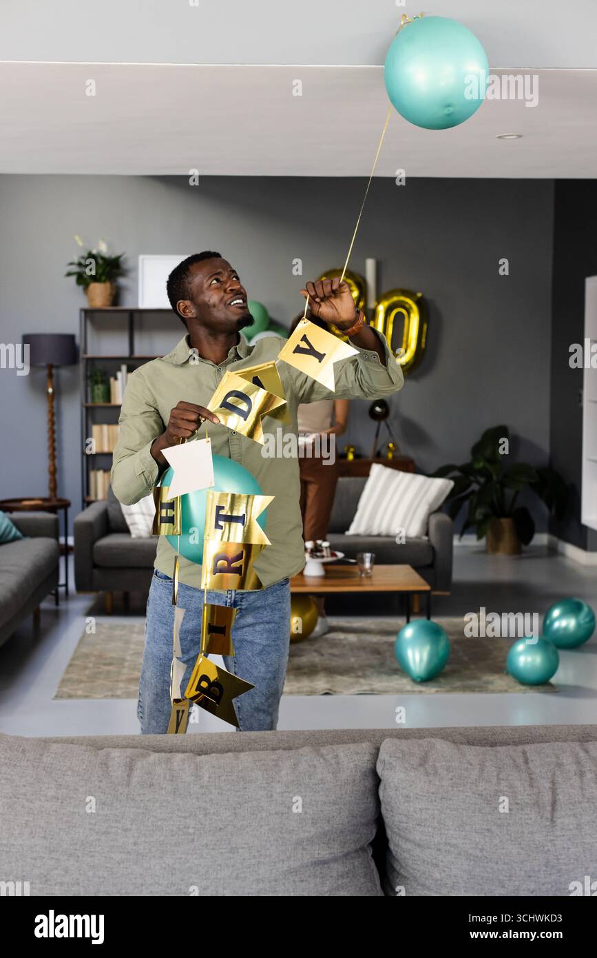 Mann dekoriert Wohnzimmer mit Geburtstagsbanner und Ballons, lächelt fröhlich Stockfoto