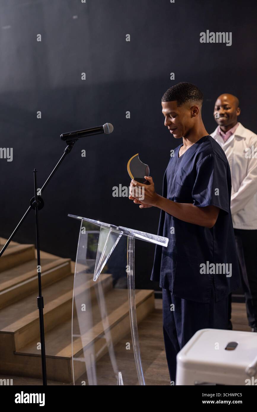 Junger Mann in Schutt, der den Preis auf dem Podium hält, lächelt mit einem Kollegen in der Nähe Stockfoto
