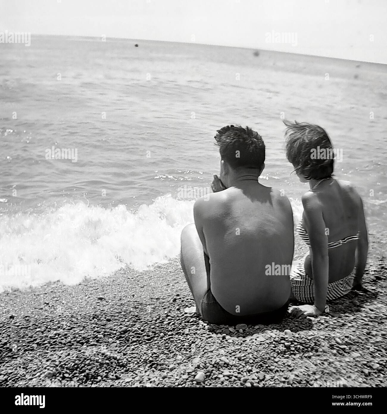Ein Archivfoto in Schwarz-weiß, aufgenommen um 1965 in Gurzuf. Ein junges Paar, ein Mann und eine Frau in Badeanzügen, sitzen zusammen an einem Kiesstrand und blicken auf den Horizont. Die Szene ist von einer romantischen, intimen und ruhigen Atmosphäre erfüllt. Das Foto fängt den Geist der „Tauzeit“ ein und ist ein Symbol für Liebe, Jugend und friedlichen Urlaub an der Krimküste in den 1960er Jahren Stockfoto