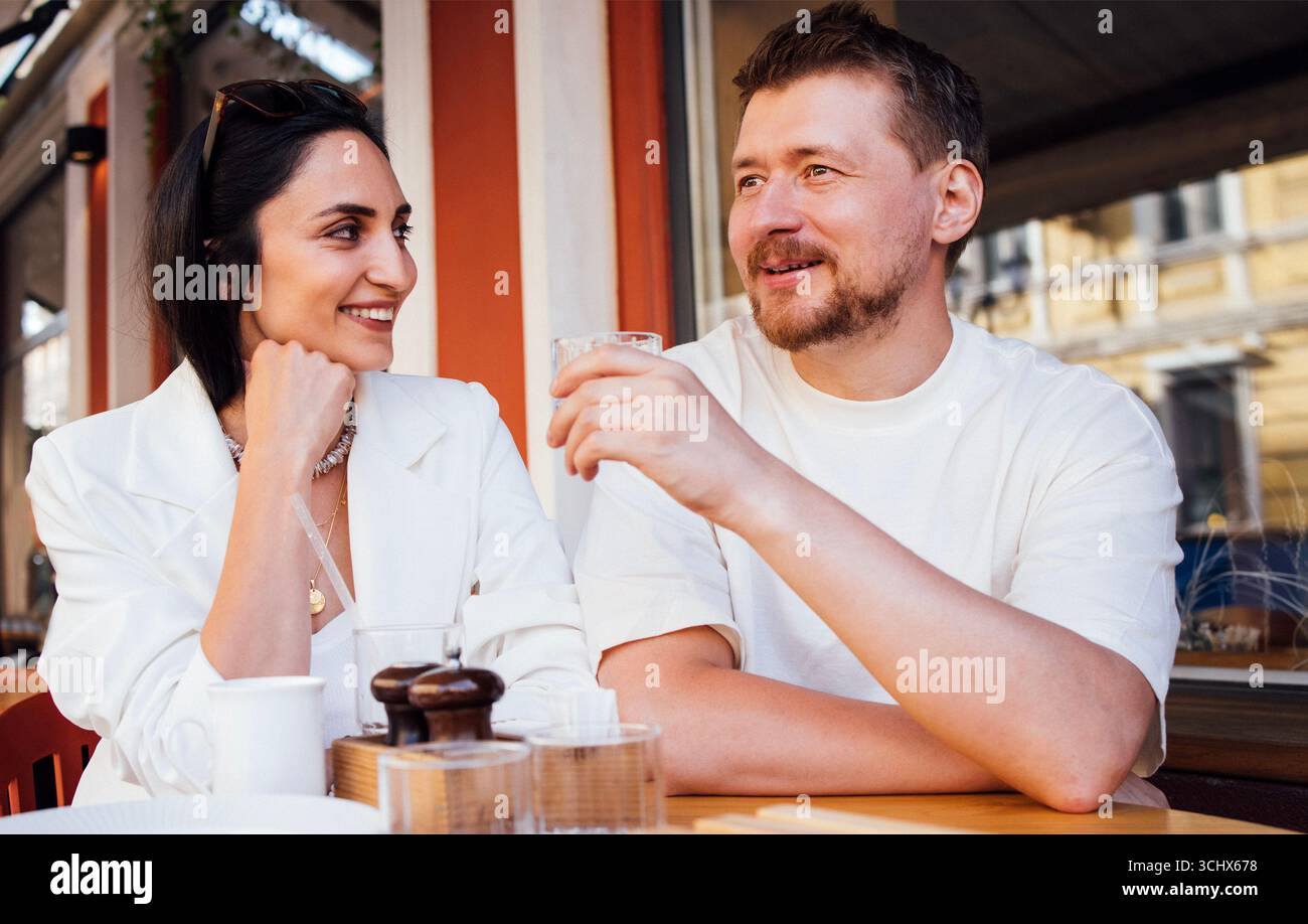 Mann und Frau sitzen an einem Café-Tisch, genießen Drinks und unterhalten sich mit einer lebhaften urbanen Kulisse, die das warme Ambiente unterstreicht Stockfoto