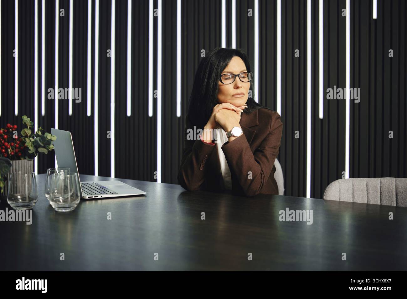 Eine professionelle Frau sitzt an einem modernen Schreibtisch in einem eleganten Bürobereich. Sie legt ihr Kinn auf die Hände, verloren in der Besinnung mit einem Laptop nahe offen Stockfoto
