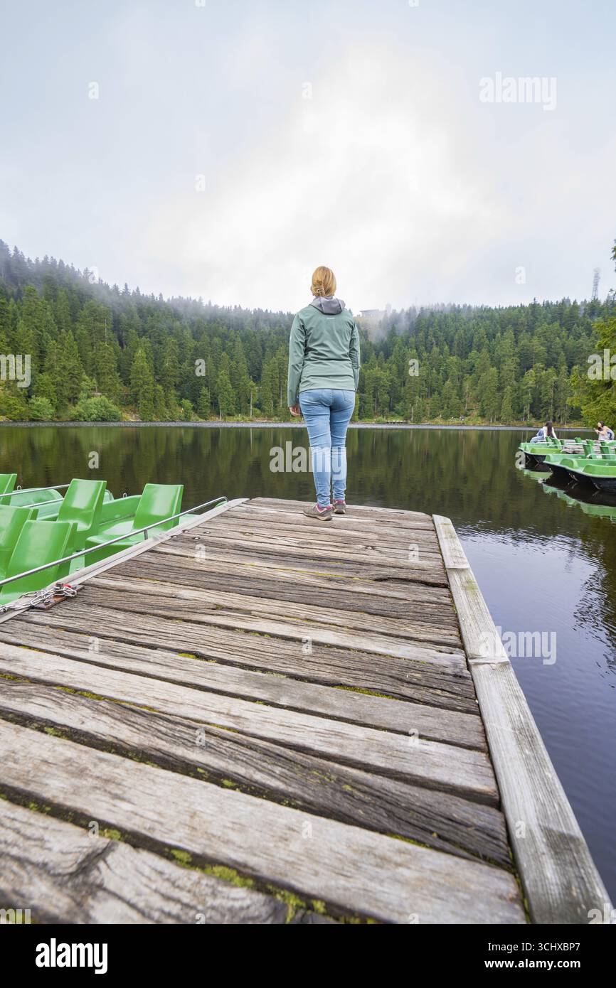 Frau, die allein auf einem Holzsteg vor einem ruhigen See steht, umgeben von Wald, Mummelsee, Seebach, Ortenau, Schwarzwald, Deutschland Stockfoto