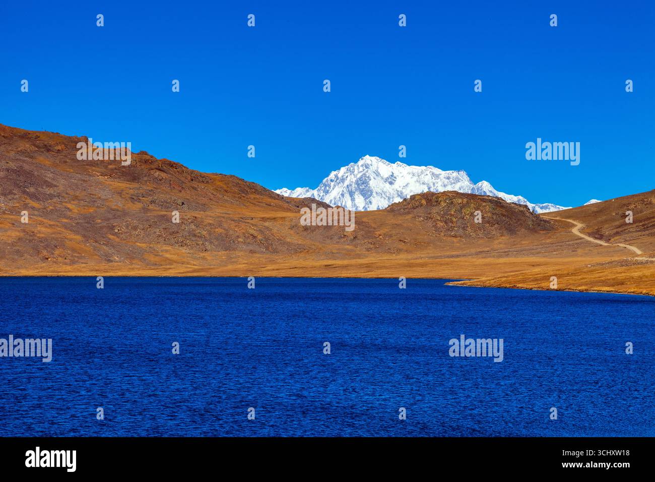 Blick auf einen ruhigen, dunkelblauen See, der den klaren azurblauen Himmel spiegelt, eingerahmt von zerklüfteten braunen Bergen und einem entfernten schneebedeckten Gipfel, Deosai Nationalpark, Gilgit Baltistan, Pakistan. Stockfoto