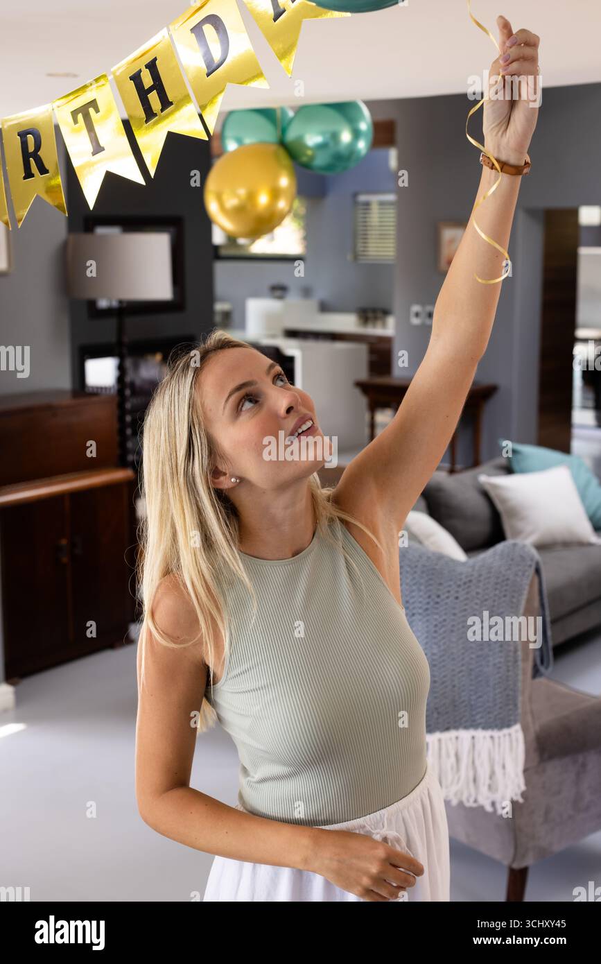 Frau dekoriert Wohnzimmer mit Geburtstagsbanner, bereitet sich auf die Party vor, zu Hause Stockfoto