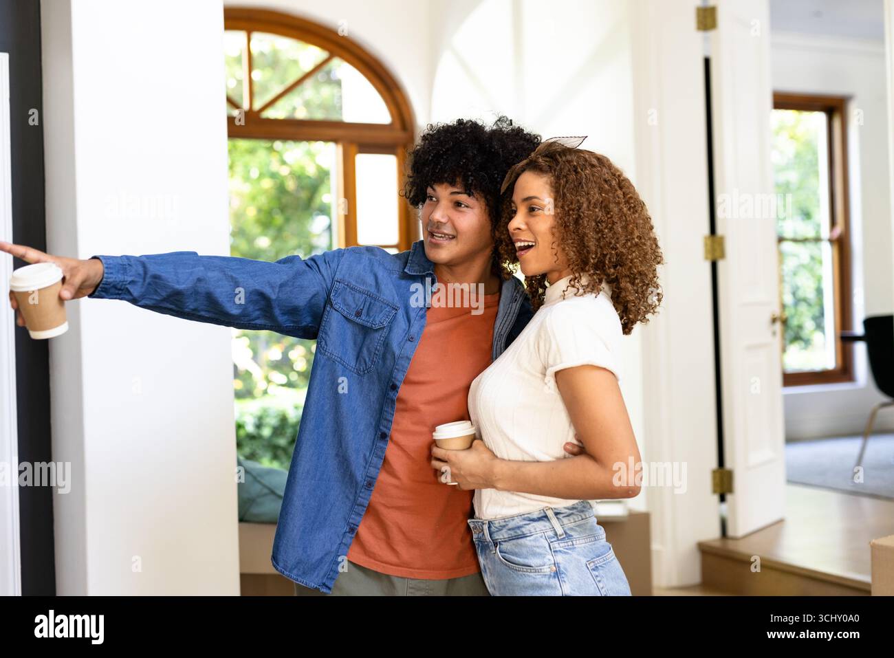 Junges, vielfältiges Paar genießt Kaffee zu Hause, lächelt und zeigt auf etwas aufregendes Stockfoto