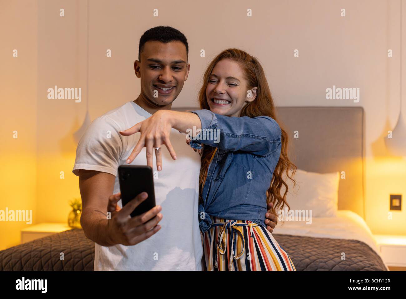 Junges vielseitiges Paar, das Verlobung feiert, Ring zeigt, Selfie im Schlafzimmer macht, zu Hause Stockfoto