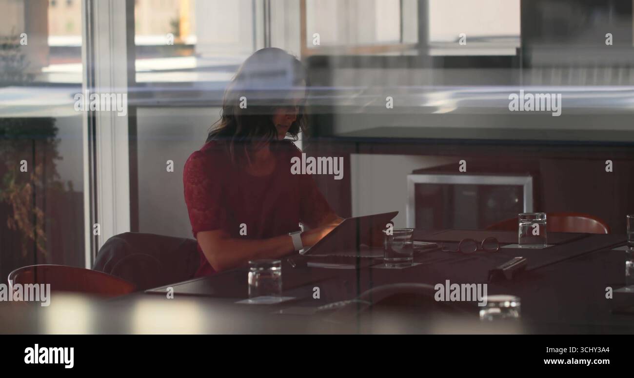 Klopfende Frau in roter Bluse, die im Konferenzraum ein Tablet mit Wasser und Lesebrille sieht Stockfoto