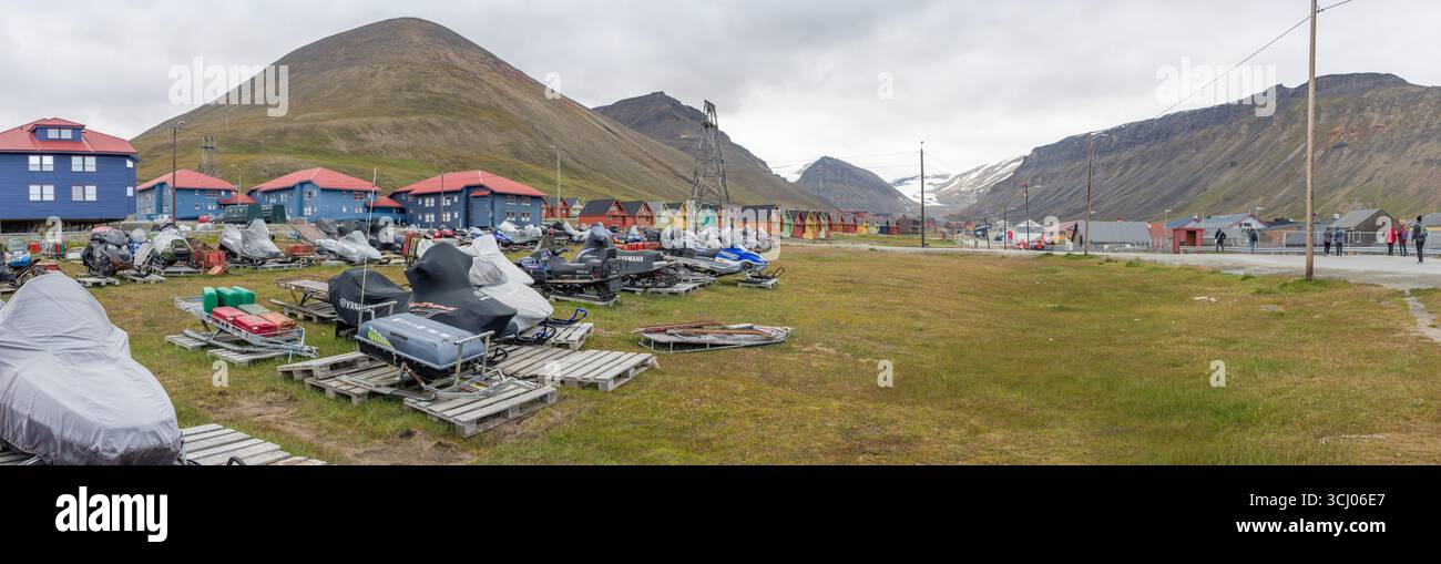 Schneemobile und Schlitten parken vor bunten Häusern Longyearbyen Svalbard Norwegen // LONGYEARBYEN, SVALBARD – Schneemobile und Schlitten parken auf einem grasbewachsenen Gelände vor bunten Häusern in Longyearbyen, dem größten Siedlungs- und Verwaltungszentrum von Svalbard, Norwegen. Die Stadt liegt eingebettet in ein Tal, mit steilen, kargen Bergen, die hinter den Gebäuden aufsteigen. Viele der Schneemobile sind im Sommer mit Schutzplanen abgedeckt. Longyearbyen ist bekannt für sein extremes Klima und ist ein Drehkreuz für arktische Forschung und Tourismus. Die unverwechselbare Architektur der Häuser, mit Stockfoto