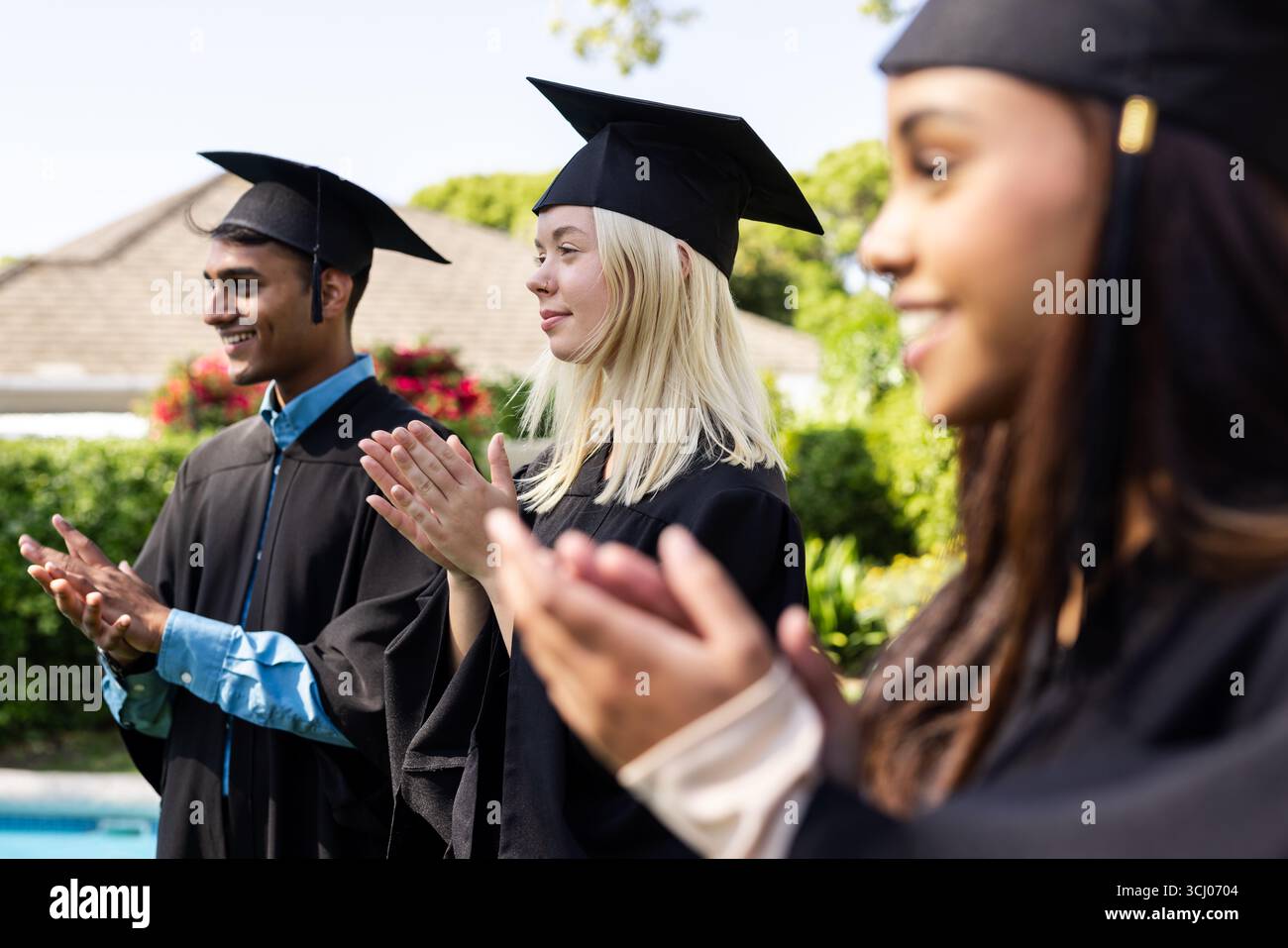 Verschiedene Absolventen in Mützen und Kleidern klatschen im Garten, um akademische Leistungen zu feiern Stockfoto