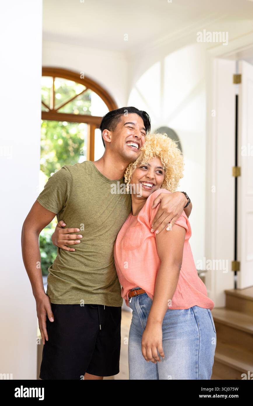Glückliches, vielseitiges Paar, das sich zu Hause in einem hellen Wohnzimmer umschließt und lacht Stockfoto