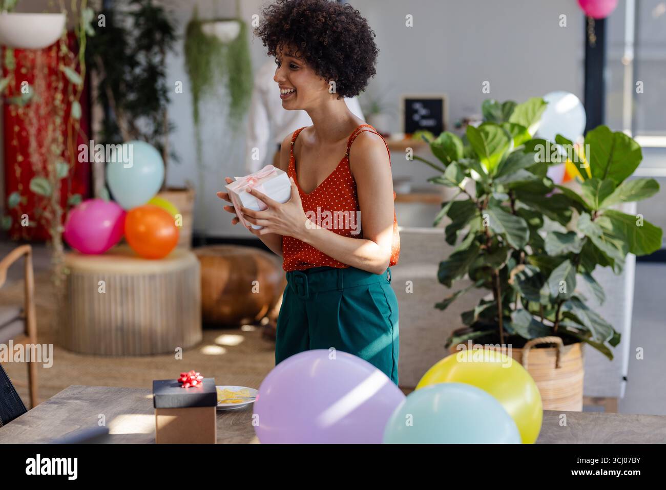 Lächelnde Frau, die ein Geschenk auf der Büroparty hält, mit bunten Ballons herum Stockfoto