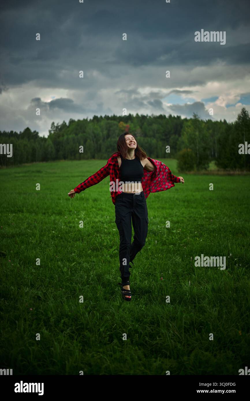 Eine Frau, die ein rotes kariertes Hemd und eine schwarze Hose trägt, wirbelt glücklich auf einem grünen Feld. Dunkle Wolken sammeln sich über sich und deuten auf wechselhaftes Wetter hin Stockfoto
