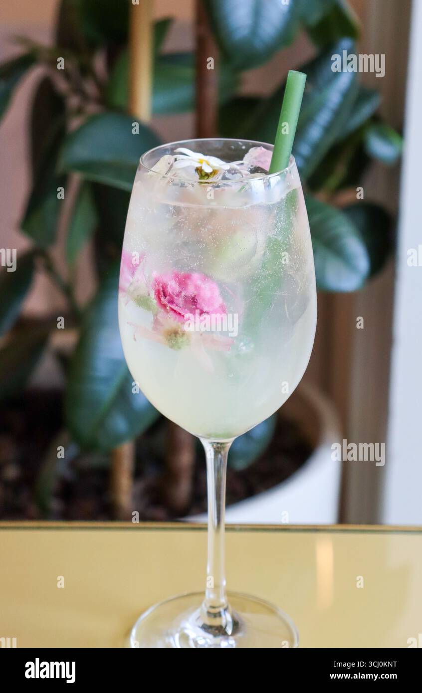 Erfrischender, floraler Cocktail in einem Weinglas mit Eiswürfeln und essbaren Blumen, garniert mit grünem Strohhalm. Prickelndes Getränk Stockfoto