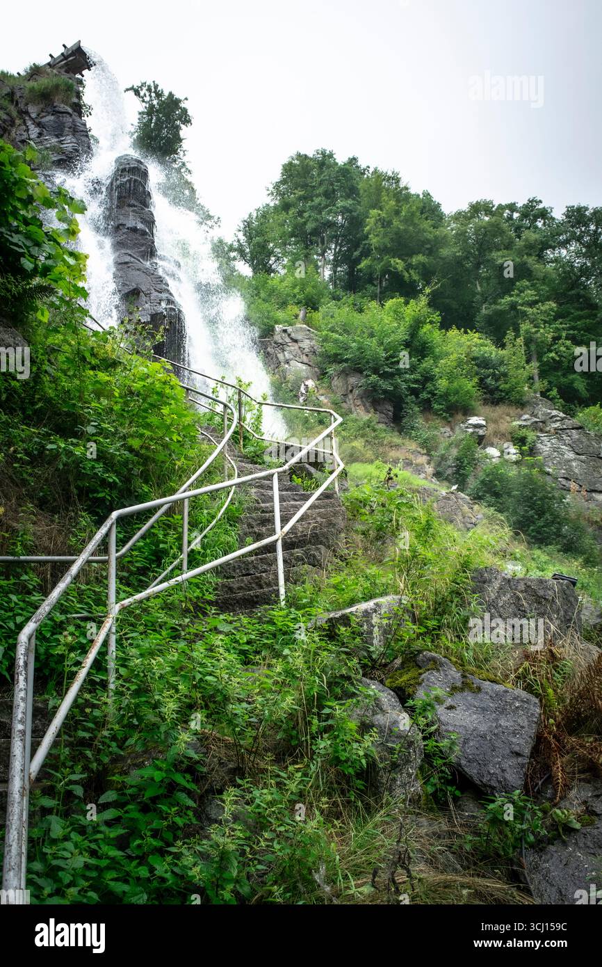 Dramatischer Wasserfall, der neben Steintreppen mit rustikalem Geländer stürzt, umgeben von üppigem Grün und Nebel, der Abenteuer und Naturwunder weckt. Stockfoto