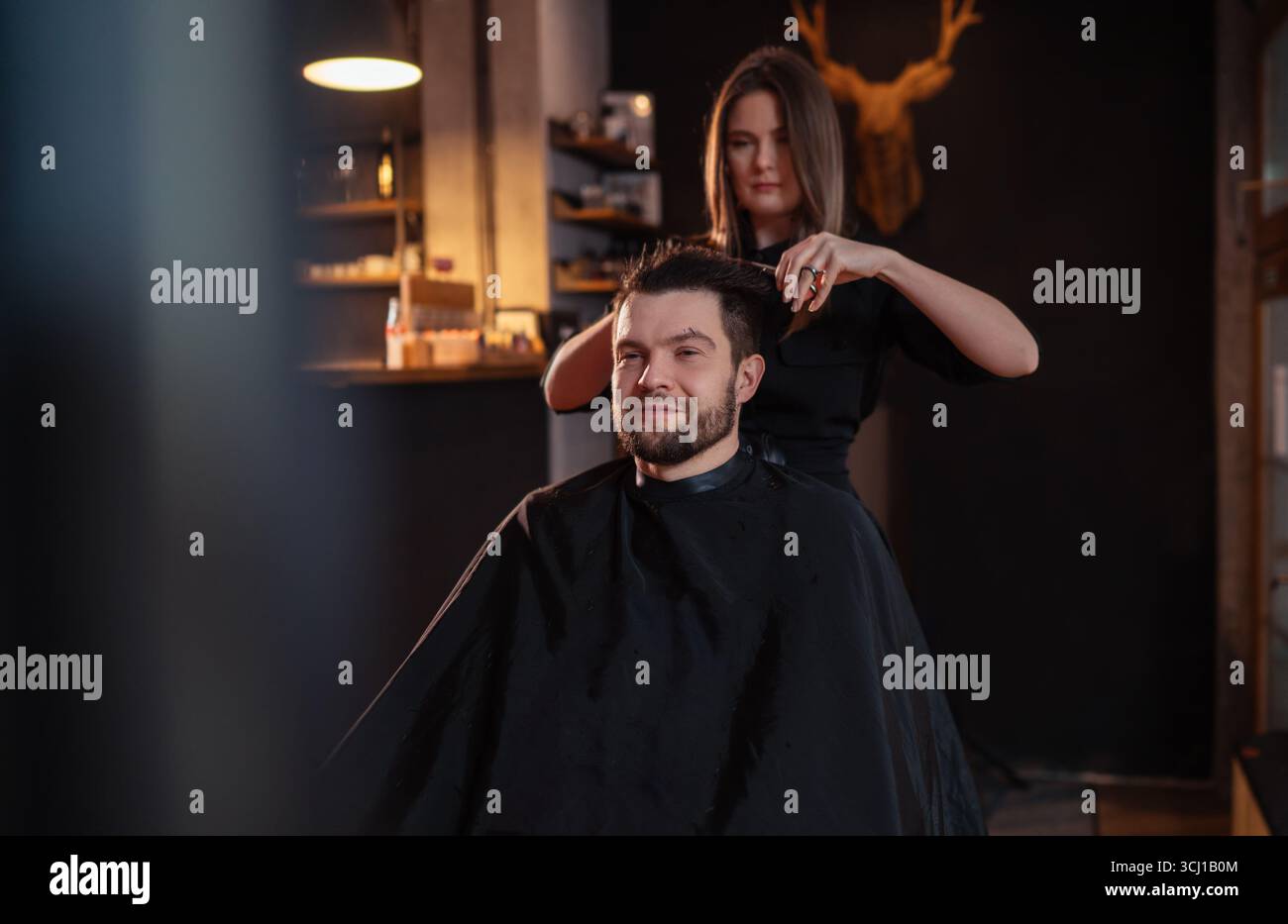 Junge langhaarige weibliche Friseurin macht sanft mit Schere und Kamm bärtigen glücklichen Mann im Salon Stuhl. Moderne Friseurstube im schwarzen Stil Stockfoto