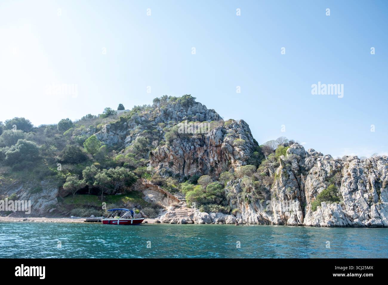 Felsige Küste mit dichtem Grün und einem kleinen Ankerboot in türkisfarbenem Wasser in der Nähe von Tolo, Griechenland. Klarer Tageslicht Himmel Stockfoto