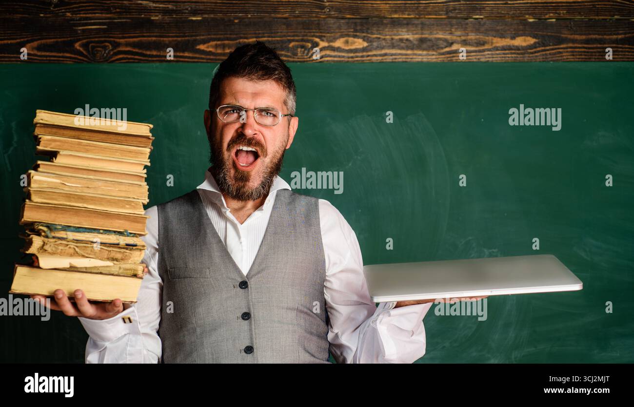 Lehrer in Brille mit Laptop und Stapel Lehrbuch im Klassenzimmer. Ernsthafter Schüler mit einem Stapel Bücher und Laptop vor der Tafel. Auswahl zwischen Modus Stockfoto