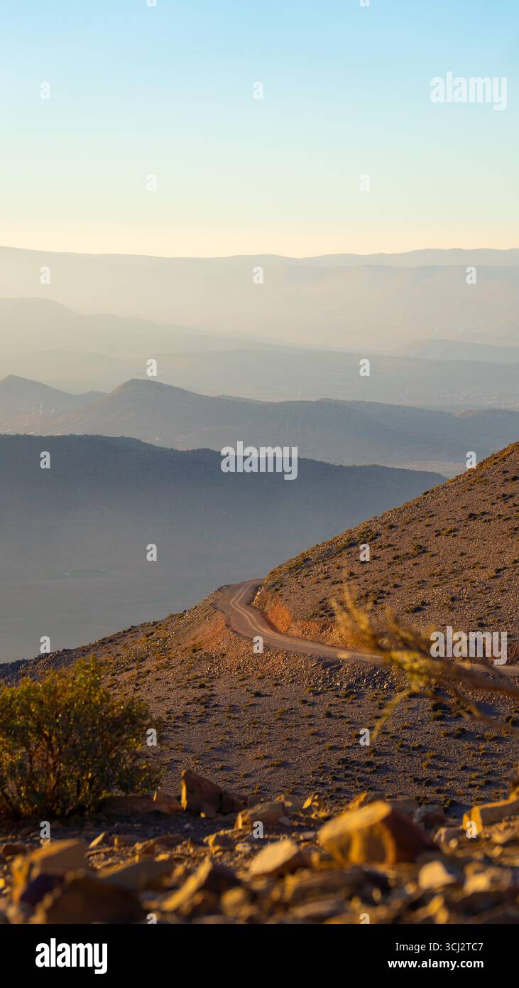 Felsiger marokkanischer Berghang mit Wüstenpflanzen und gewundenem Schotterweg mit Blick auf die neebedeckten Bergrücken unter der warmen Abendsonne Stockfoto