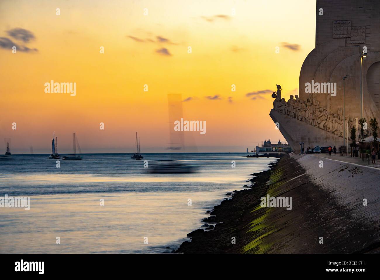 Denkmal für Entdeckungen bei Sonnenuntergang entlang des Tejo, Belém, Lissabon, Portugal mit einem Segelboot, das den Hafen vor goldenem Himmel in der Silhouette einfährt Stockfoto