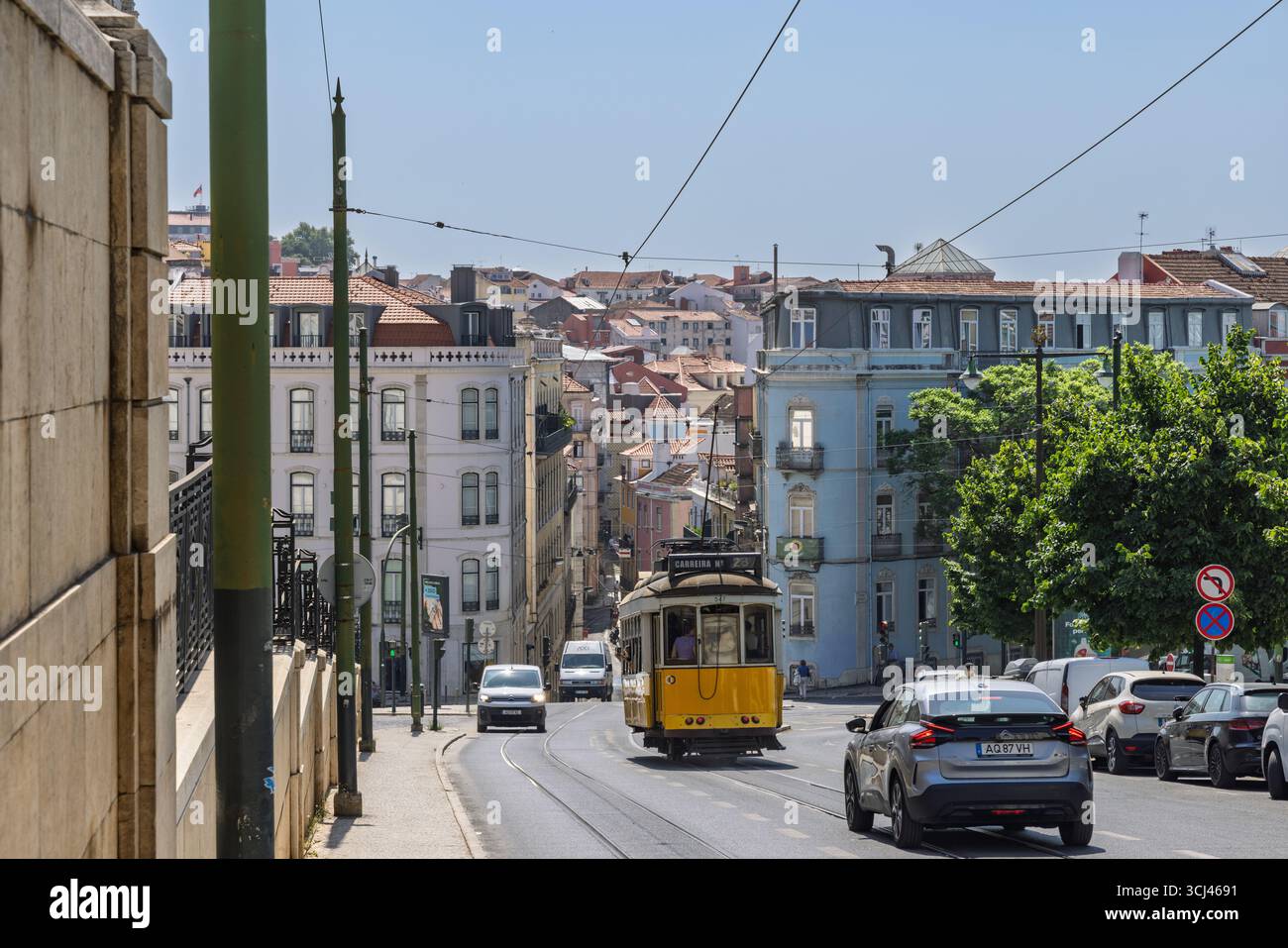 Eine klassische gelbe Lissabon-Straßenbahn fährt eine geschäftige Stadtstraße entlang, umgeben von historischen Gebäuden und der charakteristischen hügeligen Landschaft der Stadt unter einem Stockfoto