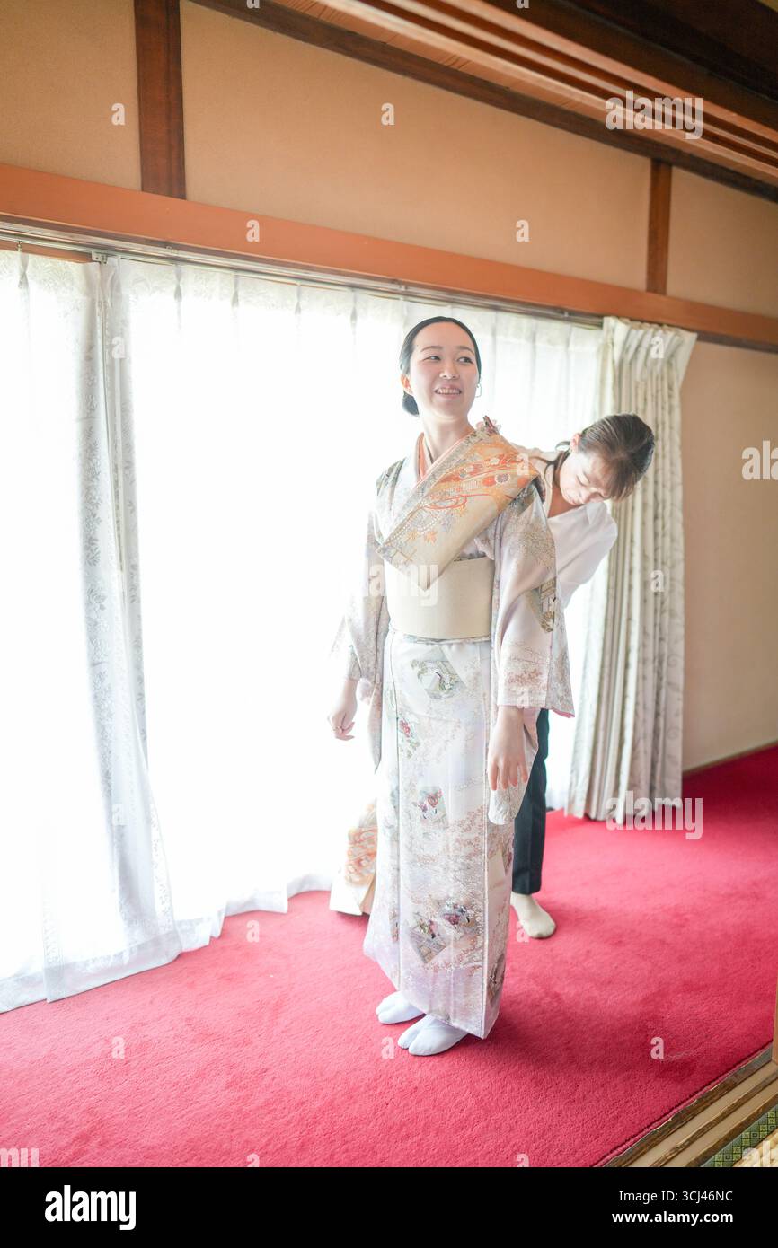 Eine Frau im Kimono zieht sich von einem Mann an. Die Frau lächelt und der Mann hilft ihr. Die Szene spielt in einem Raum mit einem Fenster und einem roten C Stockfoto