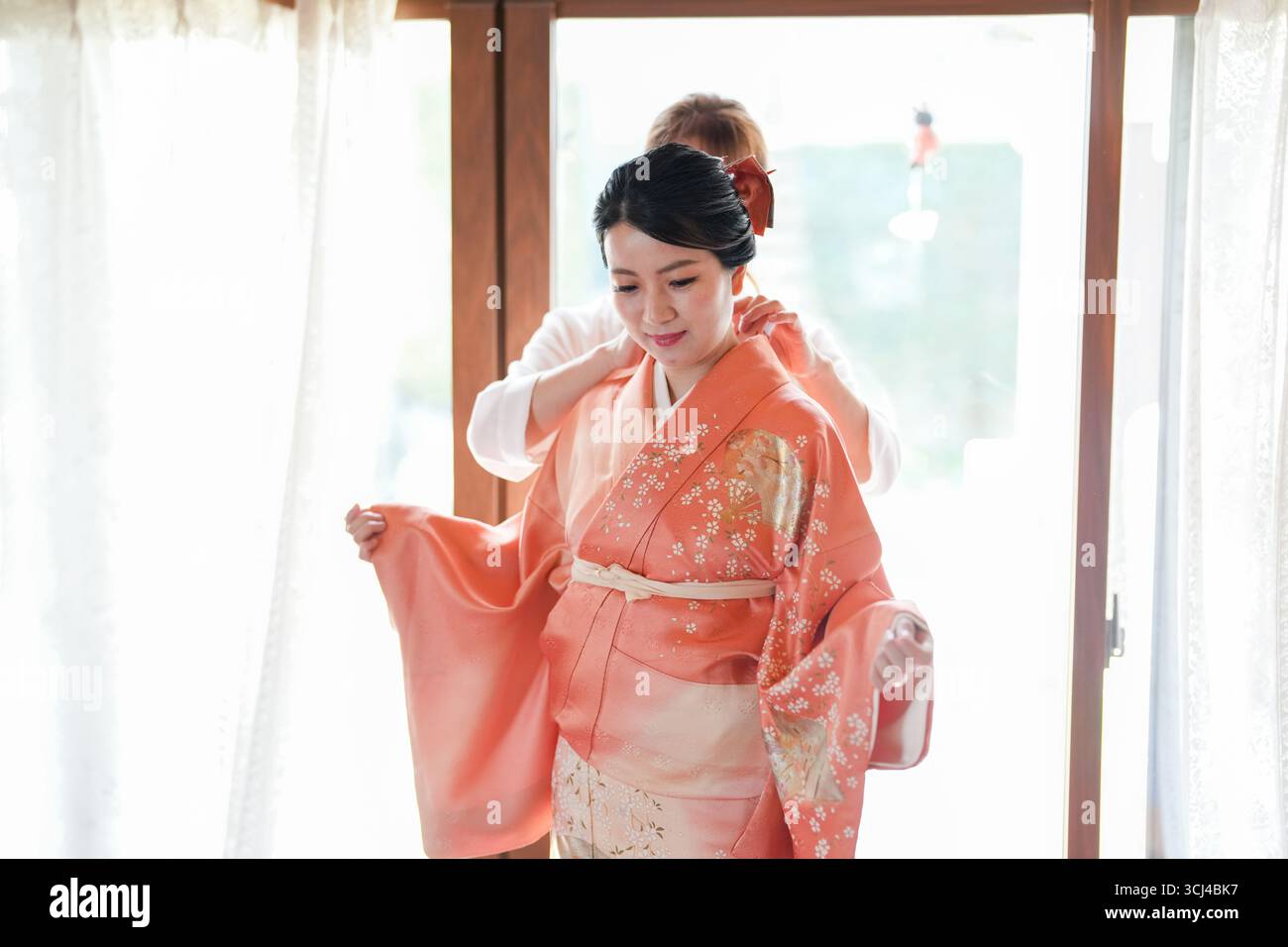 Eine Frau in einem wunderschönen orangen Kimono wird von einem Mann dabei unterstützt, ihn anzuziehen. Der Mann hilft ihr, den Kimono anzupassen und sicherzustellen, dass er richtig passt. Stockfoto