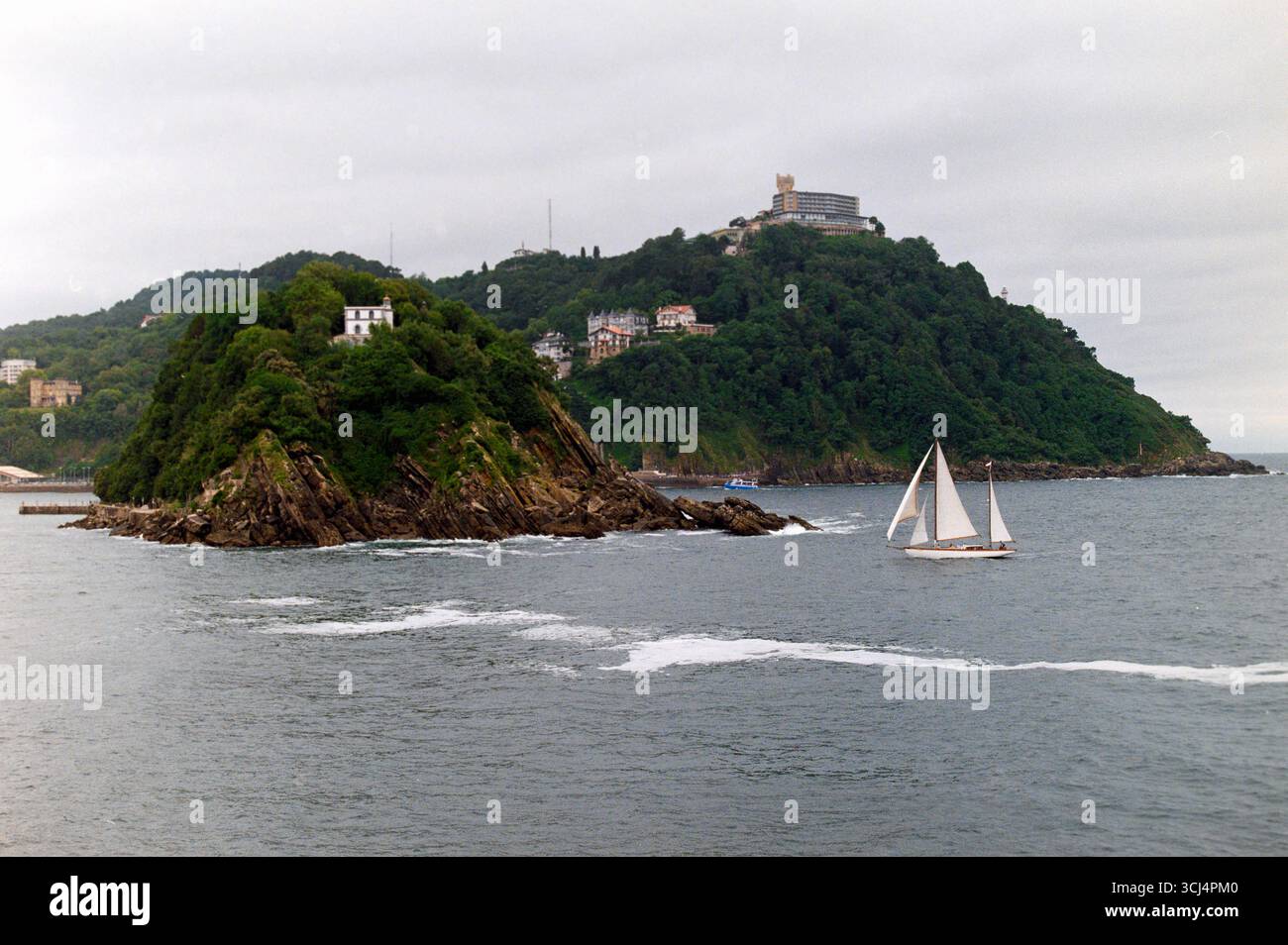 Ein Segelboot führt an einem bewölkten Tag vorbei an den üppigen grünen Hügeln von San Sebastián in Bahia de la Concha. Stockfoto