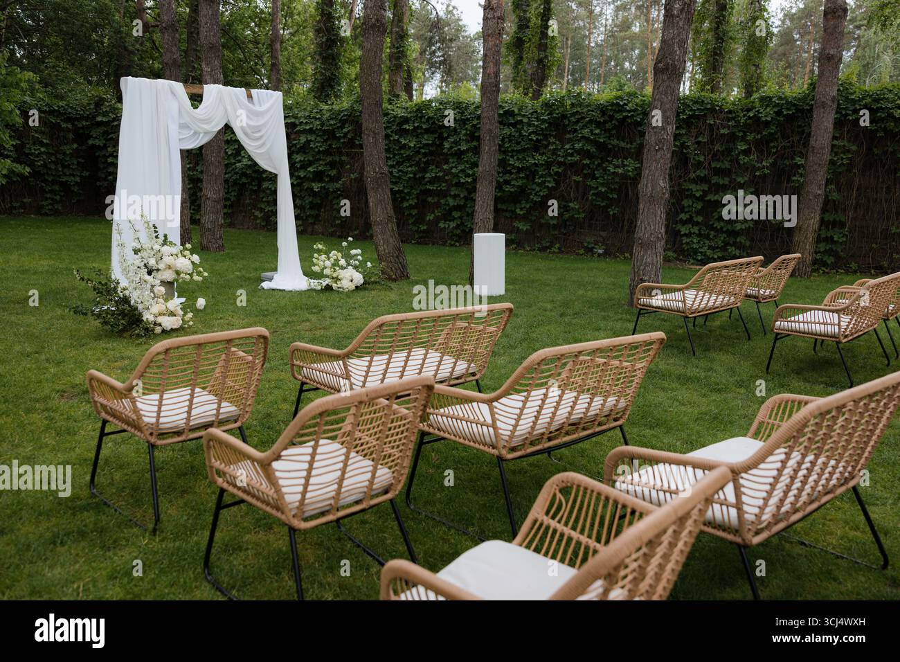 Ein bezaubernder Garten bereitet sich auf eine Hochzeitszeremonie vor. Weiße Vorhänge umrahmen den Altar elegant, umgeben von üppigem Grün und stilvollen Stühlen. Stockfoto