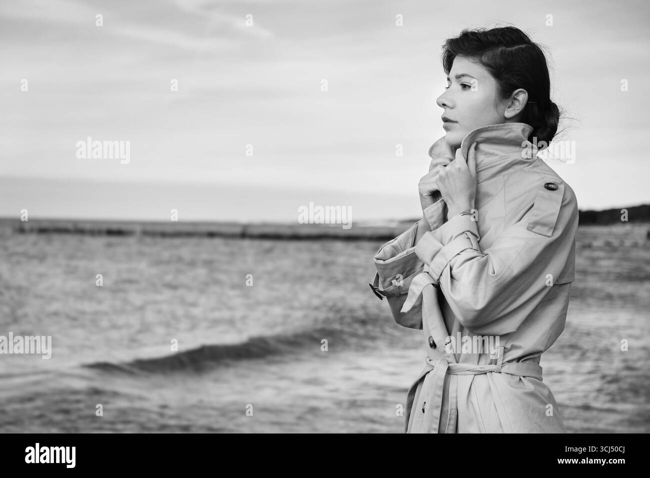 Junge Luxusfrau im Mantel in der Nähe des Meeres, die Profilansicht abschaut, monochrom Stockfoto