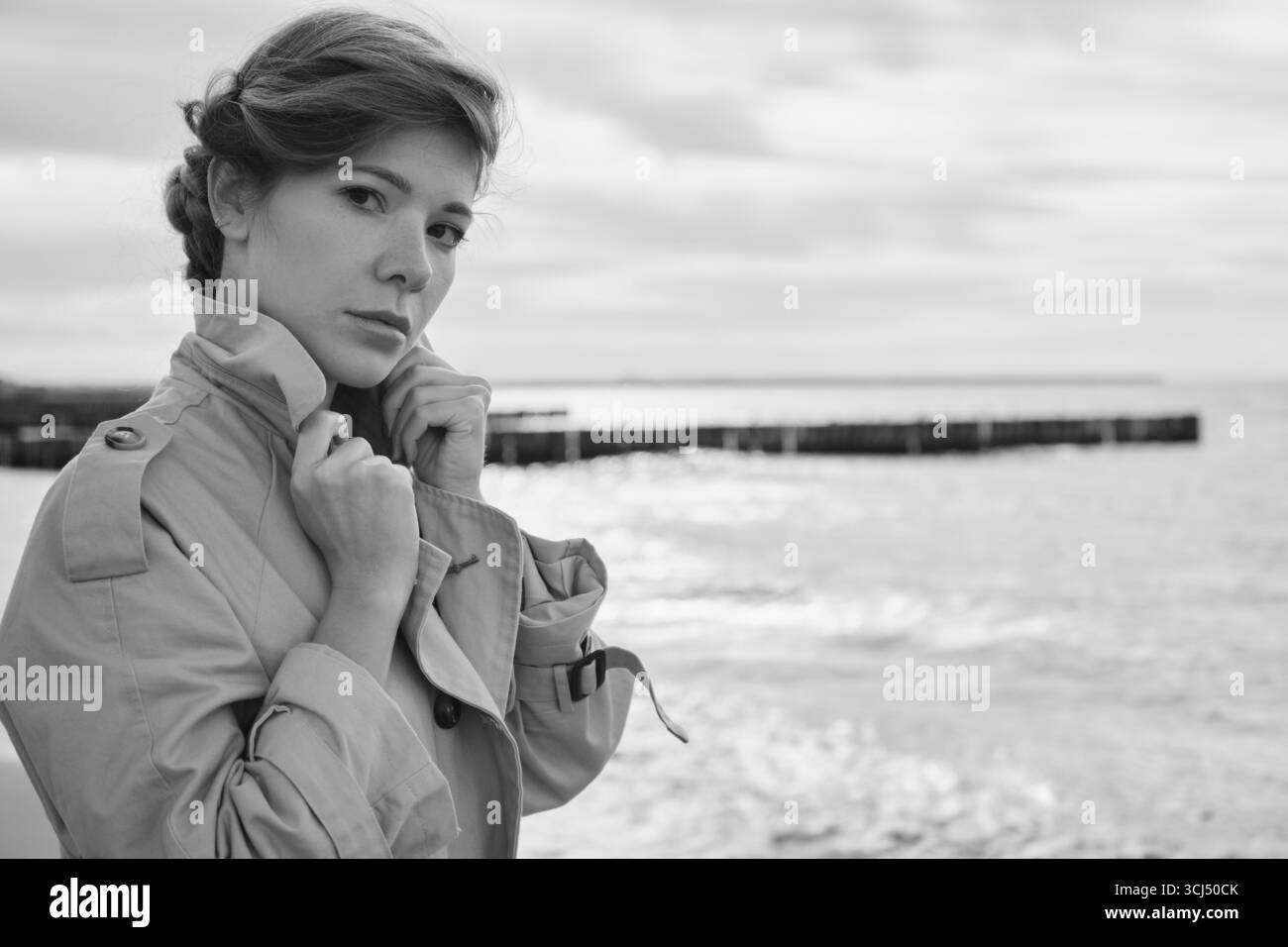 Junge Luxus-ernste Frau im Mantel in der Nähe des Meeres, die in die Kamera schaut, monochrom Stockfoto