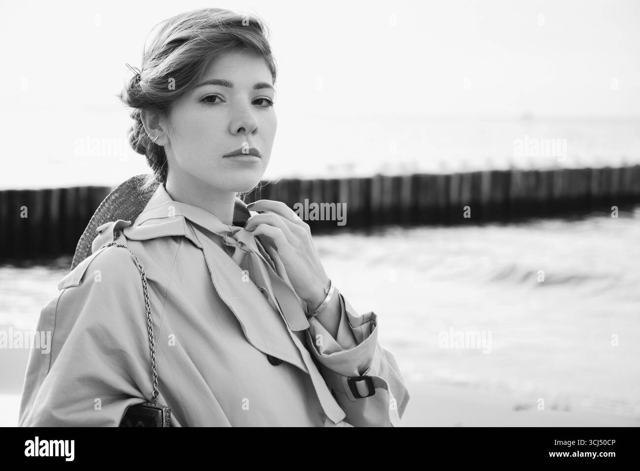 Junge Luxus-ernste Frau im Mantel in der Nähe des Meeres, die in die Kamera schaut, monochrom Stockfoto