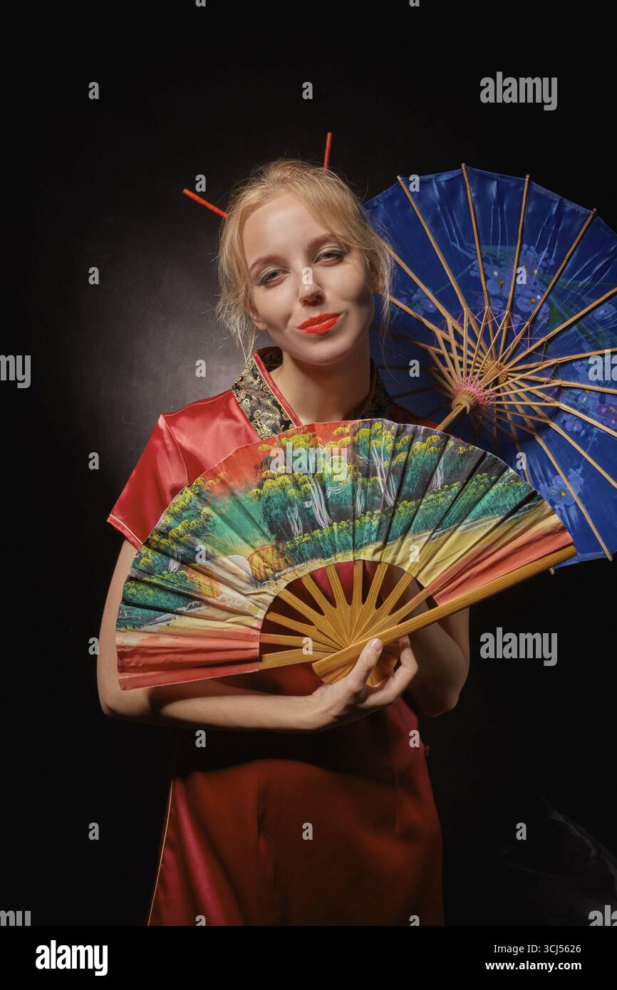 Mädchen in chinesischem Kostüm mit Fächer und Regenschirm auf schwarzem Hintergrund Stockfoto