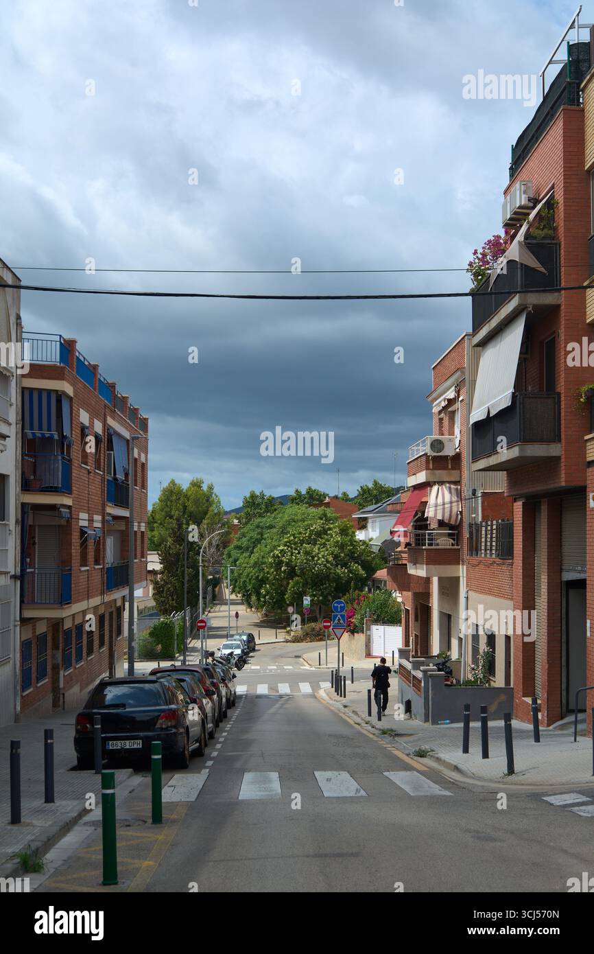 Viladecans. Barcelona - 5. September 2025: Blick nach unten auf eine steile Straße, die von Gebäuden flankiert wird. Ein dunkler, stürmischer Himmel schafft ein dramatisches, filmisches, Stockfoto