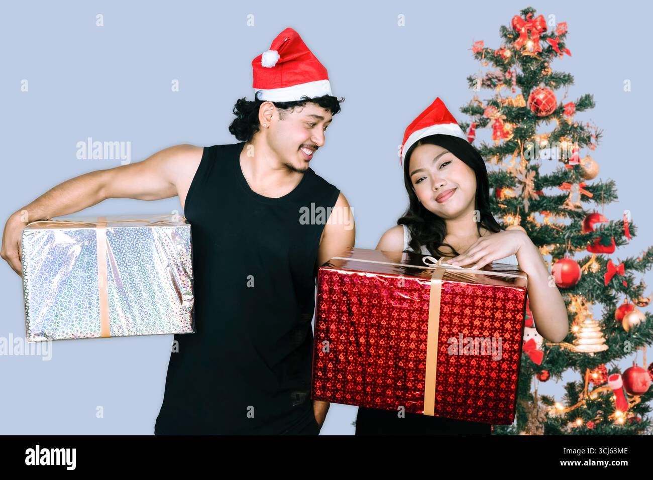 Passen Sie männliche und weibliche Athleten, die vor einem Weihnachtsbaum stehen, mit Fitnessgeräten und Geschenken, um die Weihnachtszeit zu feiern und gleichzeitig Kraft zu zeigen Stockfoto