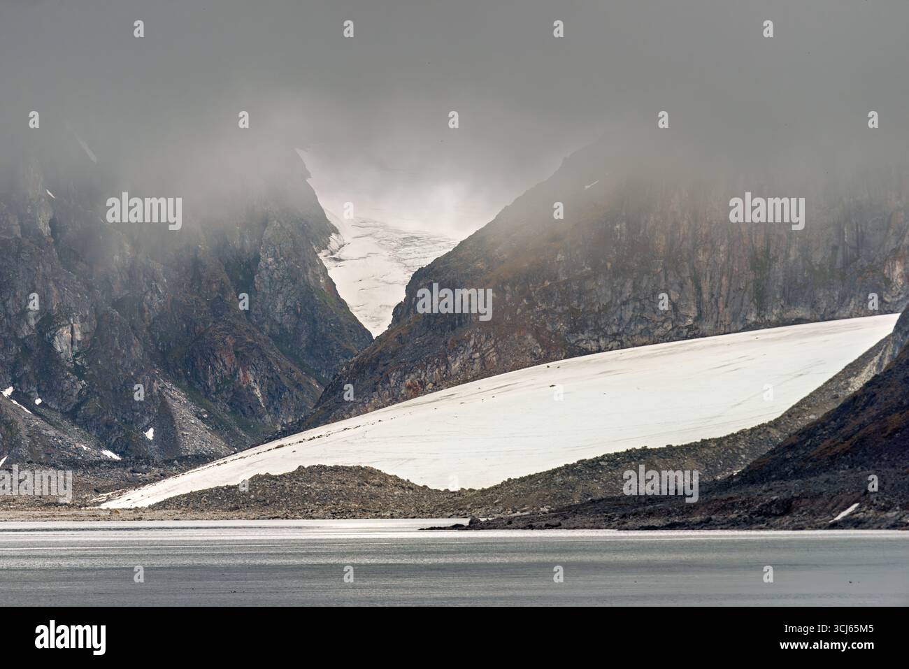 Albert I Land Glacier Fairhaven Svalbard // FAIRHAVEN, Svalbard — Albert I Land ist eine Halbinsel im Nordwesten Spitzbergens, Svalbard, Norwegen, die von schroffen Bergen und Gletschern geprägt ist. Die Landschaft besteht aus steilen, dunklen Felswänden, durchsetzt von Schnee und Eis, die zu einem Gewässer führen. Ein markanter Gletscher fließt ein Tal hinunter, teilweise von Nebel und Wolken verdeckt, was das dramatische arktische Gelände hervorhebt. Diese Region ist Teil des Svalbard-Archipels, einer arktischen Region mit hohen Breiten, die für ihre Gletscher, Berge und einzigartige Tierwelt bekannt ist. Stockfoto