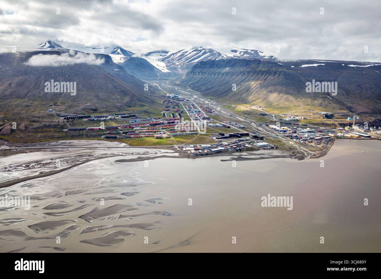 Longyearbyen Aerial View with Mountains and Fjord Svalbard Norwegen // LONGYEARBYEN, Svalbard — eine Luftaufnahme zeigt die Siedlung Longyearbyen, eingebettet zwischen steilen, schneebedeckten Bergen und einem Fjord. Die Stadt liegt auf der Insel Spitzbergen im Svalbard-Archipel und ist Norwegens nördlichste Siedlung. Die Landschaft besteht aus einem geflochtenen Flusssystem, das ins Meer fließt, mit farbenfrohen Gebäuden der Stadt, die sich entlang des Talbodens verteilen. Longyearbyen dient als Verwaltungszentrum von Svalbard und ist ein Drehkreuz für Tourismus, Forschung und Bergbau. Die umliegende Halterung Stockfoto