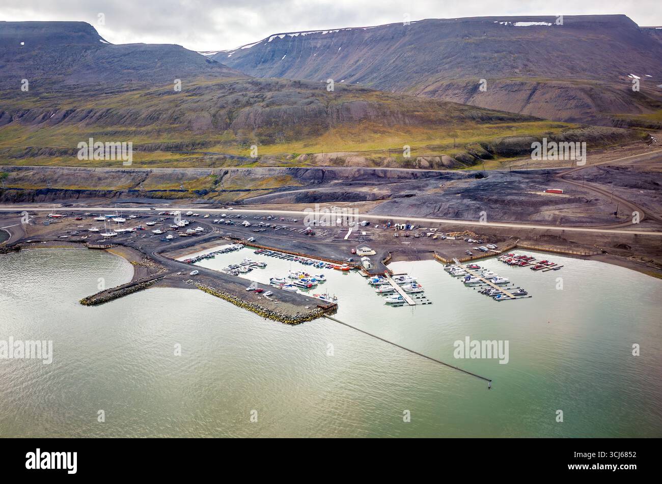 LONGYEARBYEN Svalbard Norwegen // LONGYEARBYEN, Svalbard — Ein kleiner Freizeitboothafen befindet sich an der Uferpromenade in der Nähe des Flughafens Longyearbyen, der größten Siedlungs- und Verwaltungszentrale des Svalbard-Archipels in Norwegen. Der Yachthafen verfügt über zahlreiche Boote, die entlang der Piers angedockt werden, wobei einige Schiffe auch auf dem dunklen, felsigen Gelände um den Hafen an Land gezogen werden. Im Hintergrund erheben sich zerklüftete Berge steil, deren Hänge von karger Vegetation und schneebedeckten Flecken gekennzeichnet sind. Die Landschaft ist charakteristisch für die arktische Umgebung mit einem weiten, offenen Himmel Stockfoto