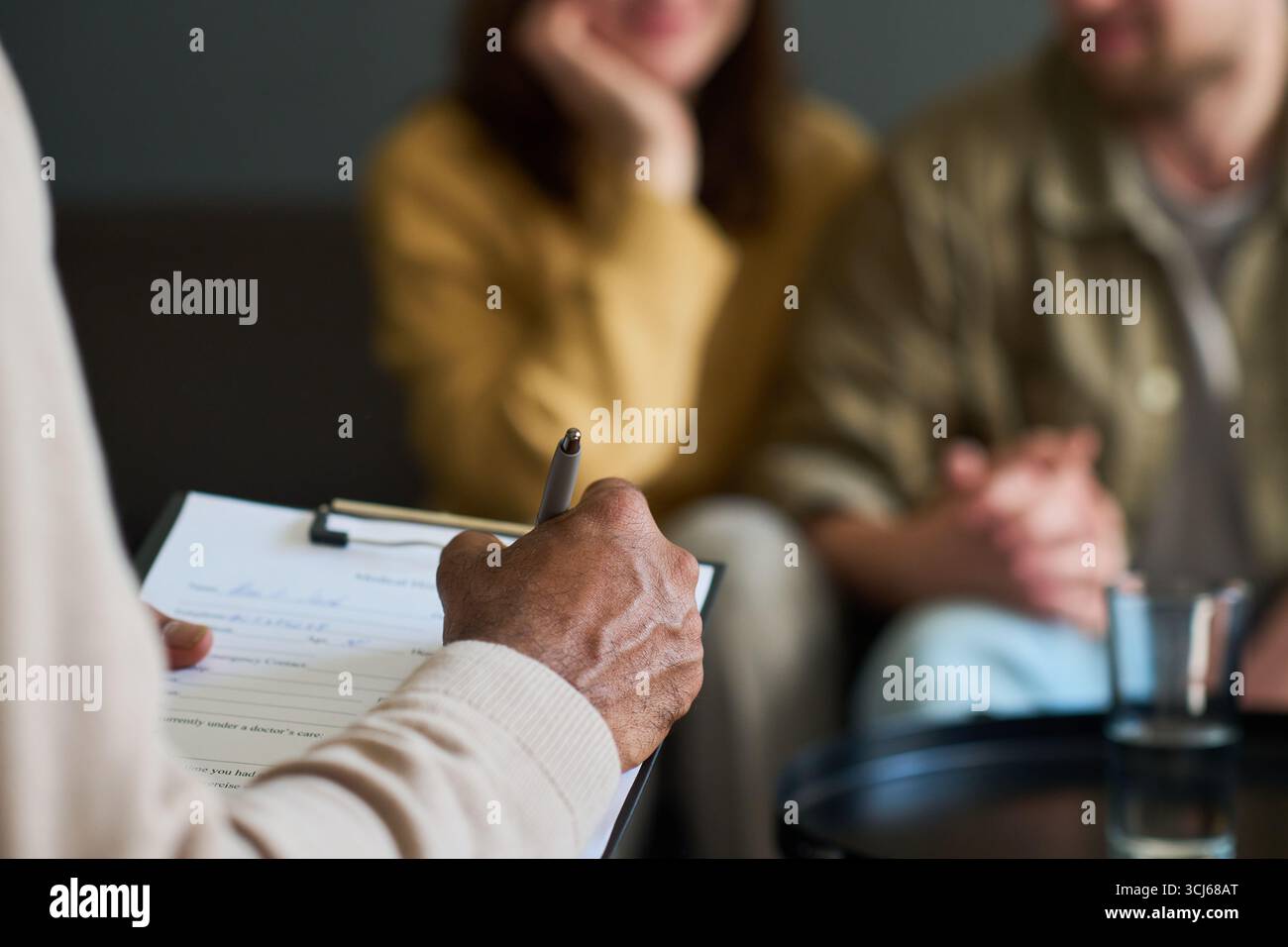 Schwarzer Mann mittleren Alters mit Klemmbrett und Stift, der eine Beratungssitzung mit jungen kaukasischen Paaren führt, die im Hintergrund zusammen sitzen, die Hände halten und aufmerksam zuhören Stockfoto