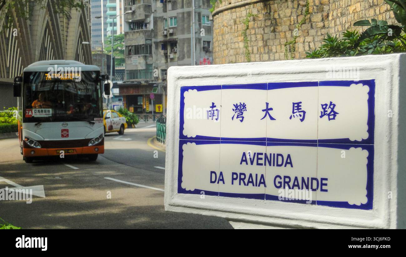 Öffentliche Busse fahren entlang der Avenida da Praia Grande in Macau, mit Portugiesisch-chinesischem Straßenschild und Stadtbild im Hintergrund. Stockfoto