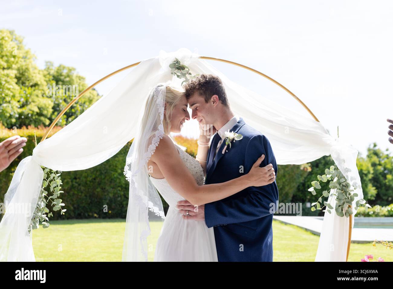 Braut und Bräutigam umschmeicheln sich unter dem Blumenbogen und teilen einen freudigen Moment im Freien Stockfoto