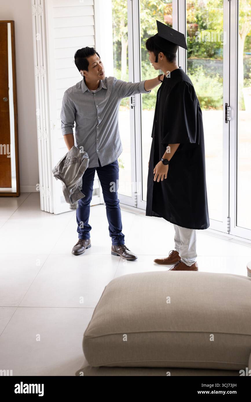 Vater gratuliert dem Sohn im Abschlusskleid zu Hause und teilt gemeinsam stolzen Moment Stockfoto