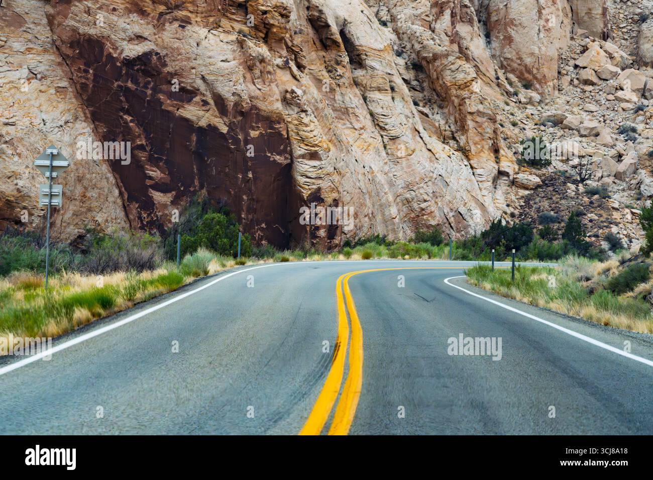 Eine leere Asphaltstraße biegt sanft nach rechts ab und führt in eine weite, zerklüftete Wüstenlandschaft. Helle, doppelte gelbe Linien markieren deutlich die Mitte von Stockfoto