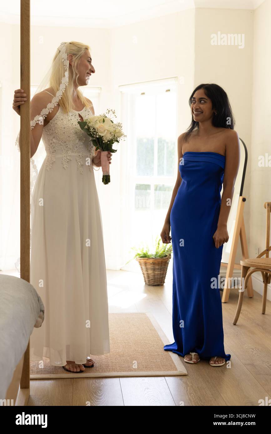 Braut mit Blumenstrauß, lächelnd mit Brautjungfer in blauem Kleid in hellem Zimmer Stockfoto