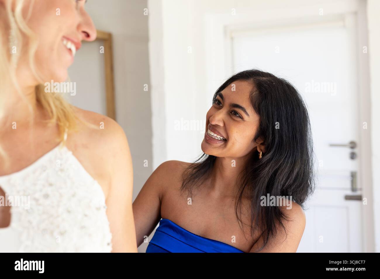 Lächelnde Frau in blauem Kleid, die Braut mit Hochzeitskleid zu Hause hilft Stockfoto