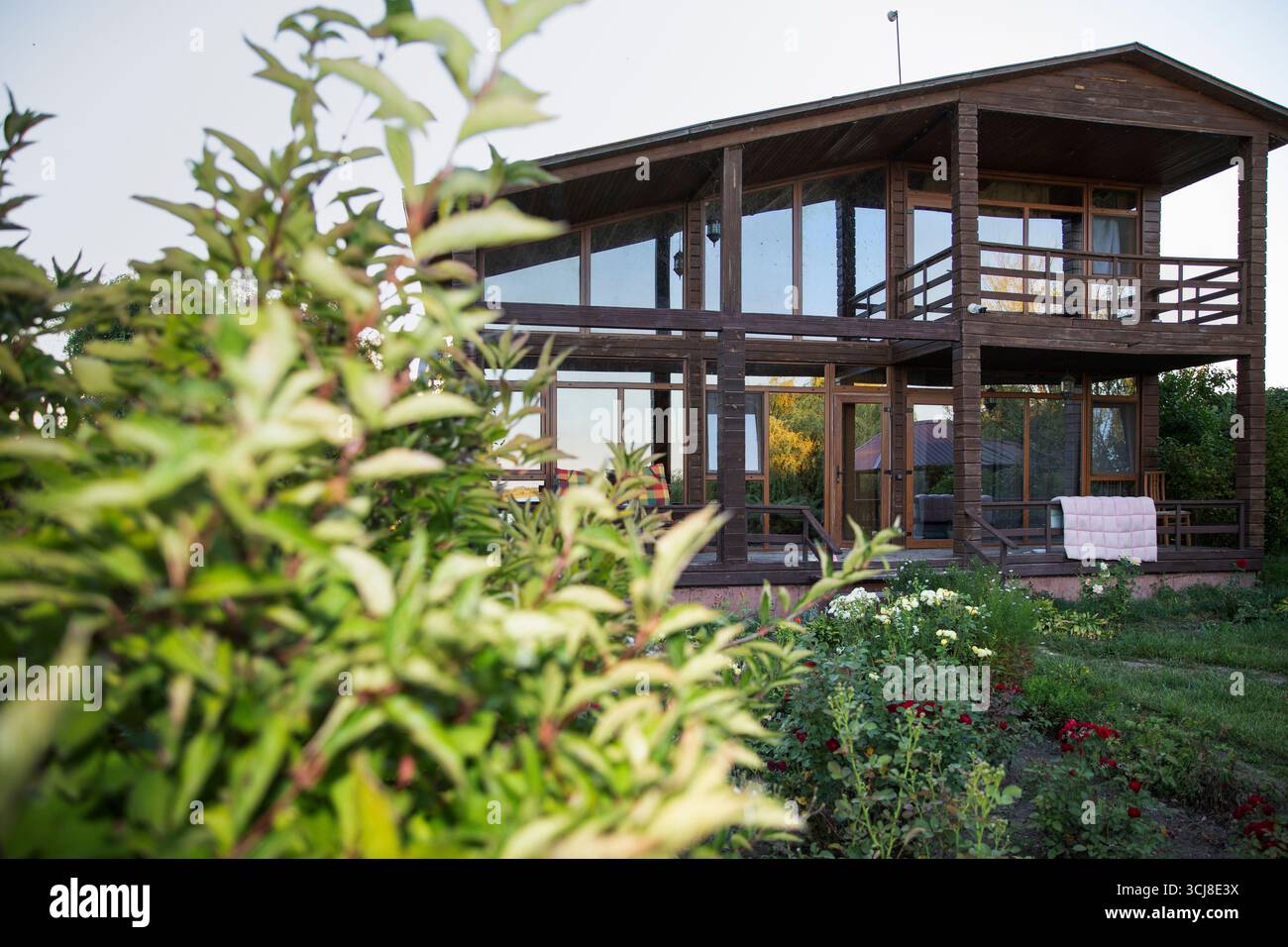 Modernes zweistöckiges hölzernes Haus mit großen Fenstern, umgeben von einem grünen Garten und Blumen schaffen eine gemütliche ländliche Wohnatmosphäre Stockfoto